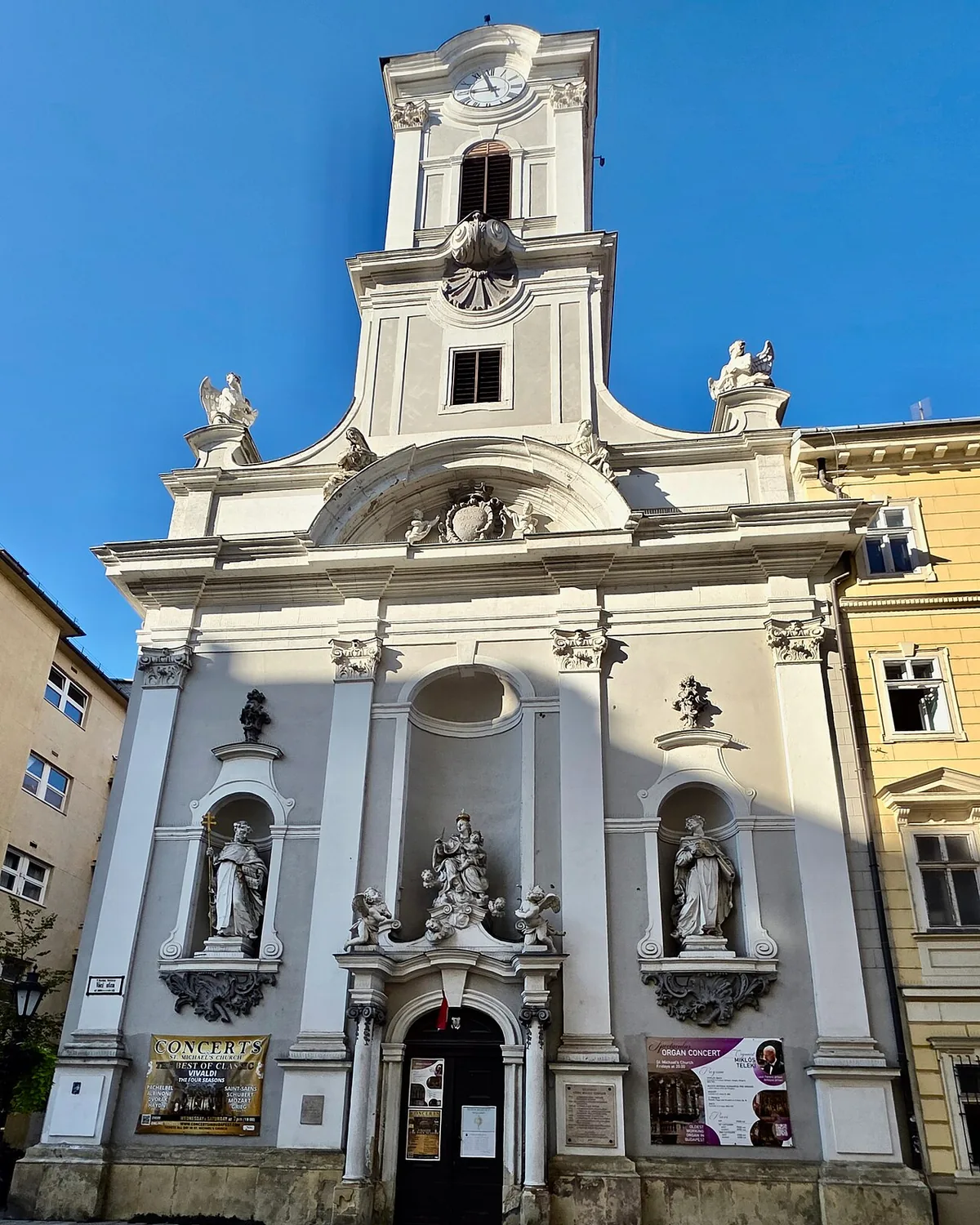The white Baroque facade of the church on Váci utca in Budapest, with a clock tower, statues in niches and a pair of columns beside the doorway under a blue sky