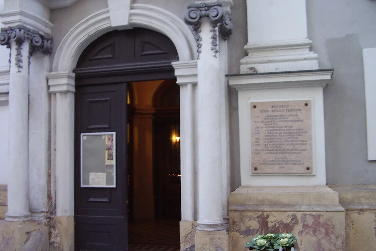 The arched stone doorway of a church on a Budapest street, with columns on both sides, a notice board and a stone plaque