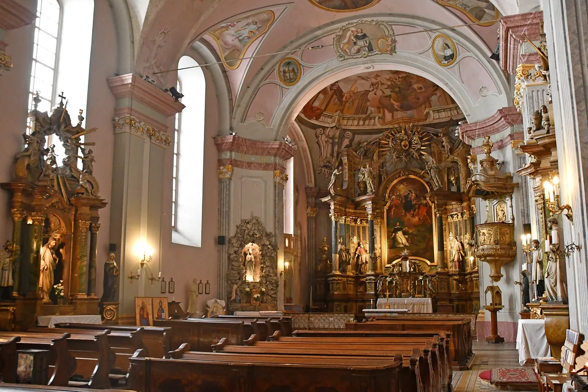 The nave of a Baroque church in Budapest, with rows of dark wooden pews facing a gilded high altar under pink-painted vaults with frescoes