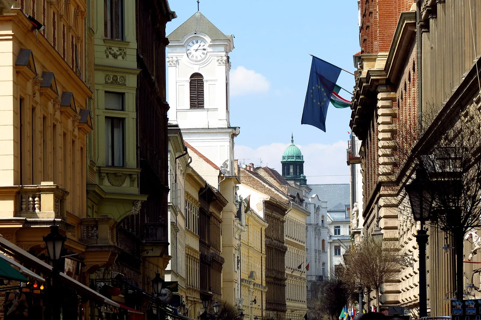 Váci utca looking towards the church, a narrow pedestrian street between tall nineteenth-century buildings, with the white clock tower rising ahead and an EU flag overhead